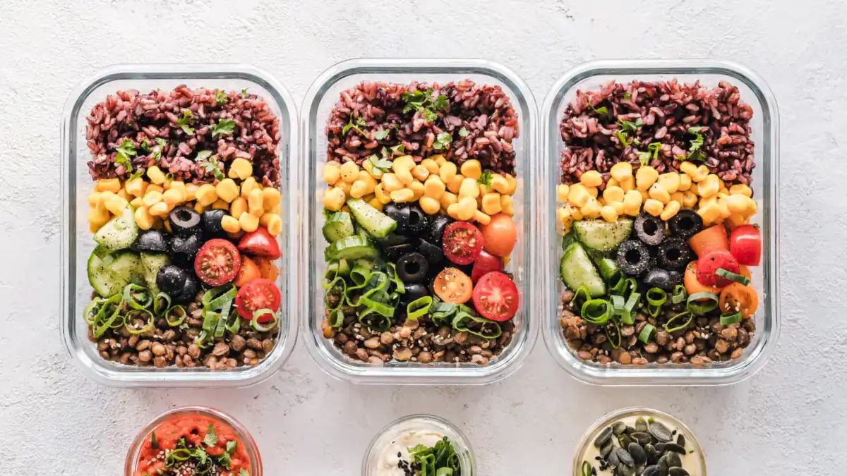 Three glass meal prep containers packed with rice, corn, olives, tomatoes and lentils, with small jars of sauce and seeds