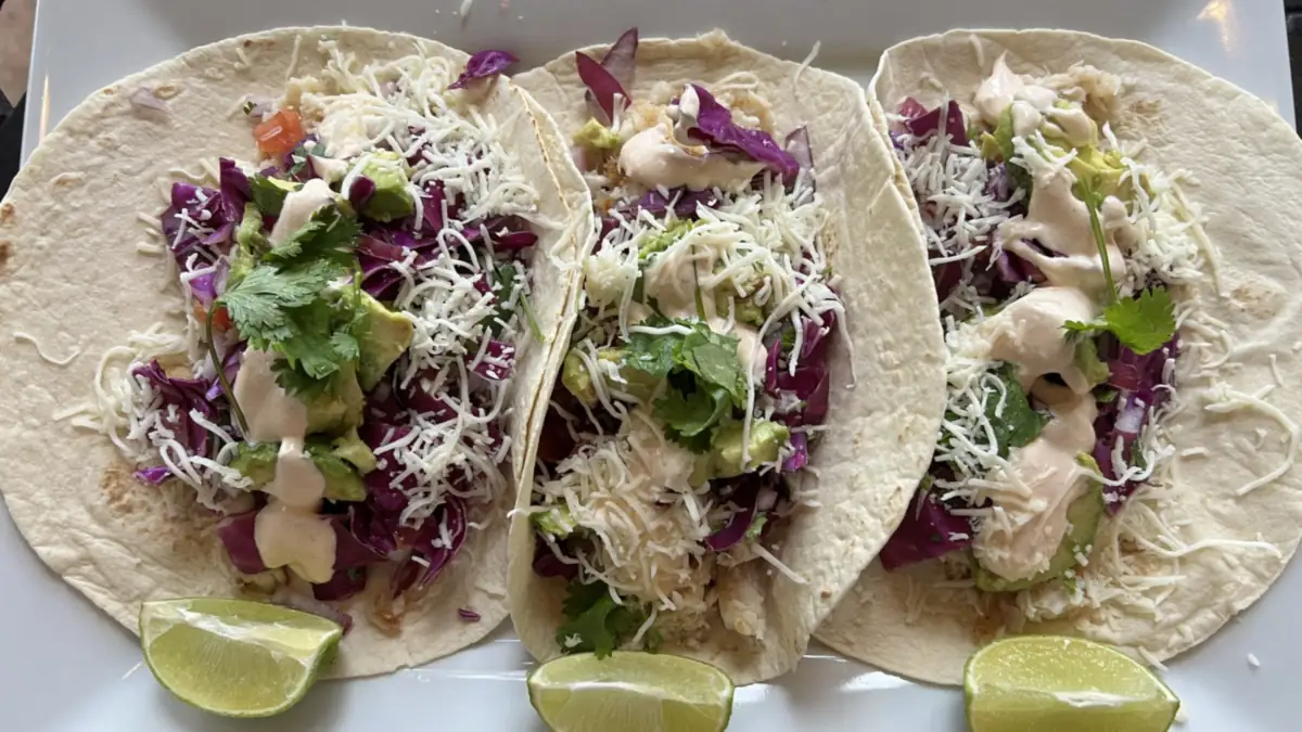 Three soft tacos topped with shredded cabbage, cheese and sauce, served with lime wedges